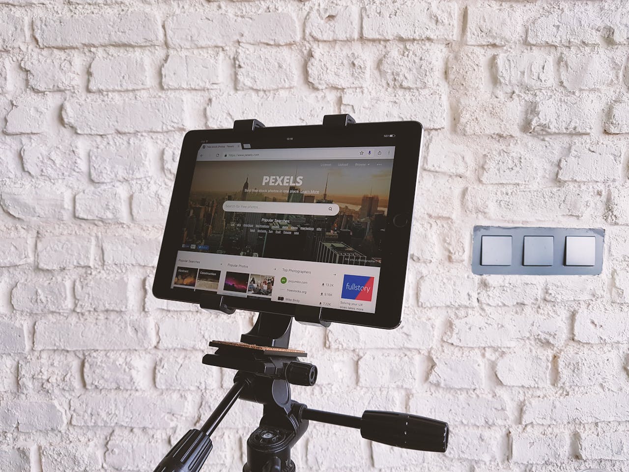 heros-img Tablet mounted on a tripod, displaying a website, set against a white brick wall indoors.